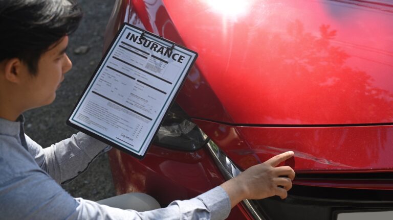 An insurance adjuster is holding a clipboard with an "INSURANCE" form, examining front-end damage on a red vehicle after a collision.